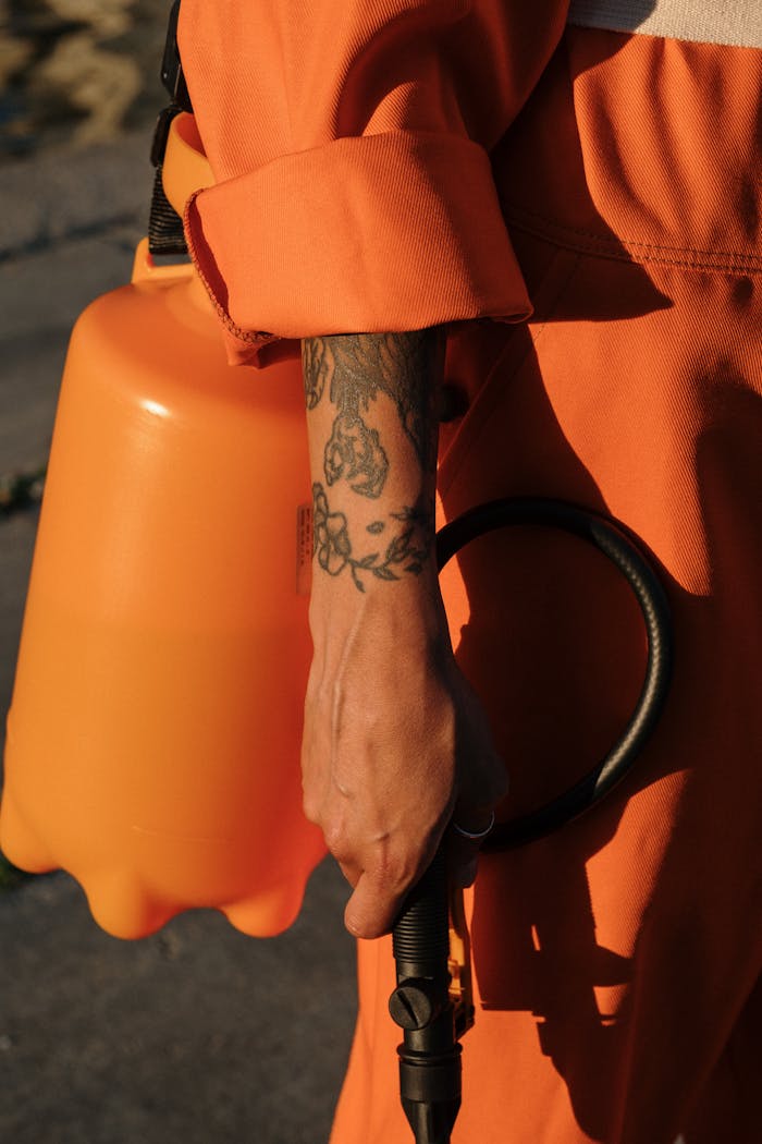 Person in orange coveralls holding a plastic sprayer, focus on tattooed arm.